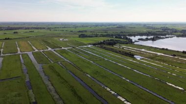 Su kanallarıyla bölünmüş uzun yeşil şeritli polder tarlalarının havadan görünüşü. Arka planda bir göl ve dağınık binalar kırsal alanda etkin arazi ve su yönetimini vurguluyor.