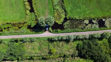 İki su gövdesi arasında çapraz bir yol var. Biri yeşillikle çevrili, biri beyaz bir araba park etmiş, biri de yol kenarında yürüyen kırmızı elbiseli bir adam..