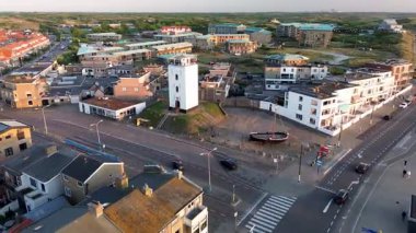 Merkezi beyaz deniz feneri, plaj ve kum tepeleri, lowrise binaları, tekne anıtı olan küçük bir plaza ve kıyı şeridinde organize bir karayolu ağı olan bir sahil kasabasının havadan görüntüsü..