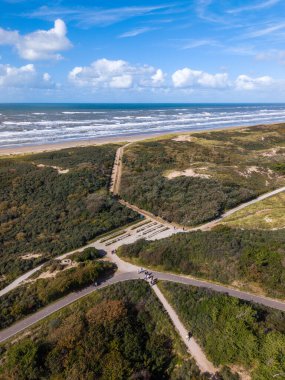 Aerial view of a bright coastal scene with a sandy beach, white capped waves, green dunes, and intersecting walking paths, dotted with people under a mostly clear sky.