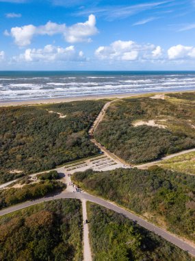 Aerial view of a bright coastal scene with a sandy beach, white capped waves, green dunes, and intersecting walking paths, dotted with people under a mostly clear sky.
