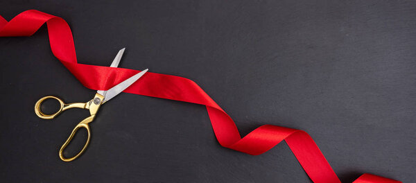 Grand opening. Top view of gold scissors cutting red silk ribbon against black background, banner, copy space.