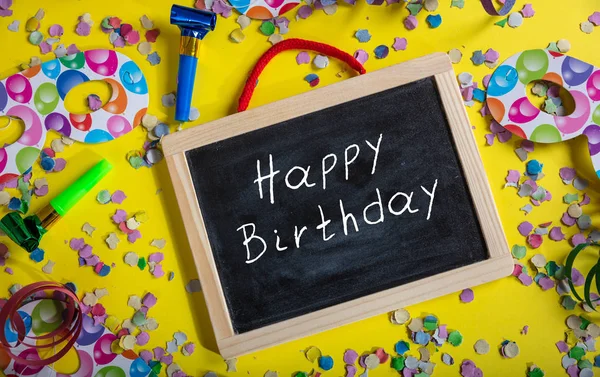 Birthday party. Happy birthday text on a blackboard, confetti and serpentines on bright yellow background, top view