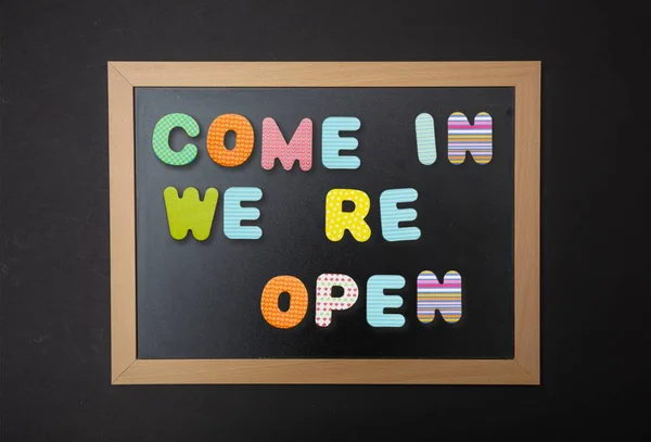Open, come in. Board with black frame, text Come in, we re open in colorful letters, black wall background