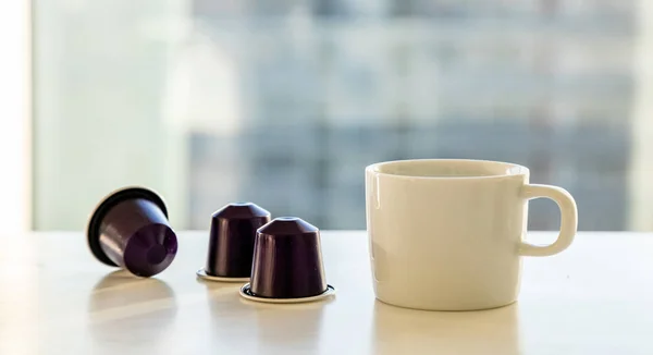 흰색 테이블에 에스프레소 커피 컵과 커피 캡슐. 흐림 창 배경 — 스톡 사진
