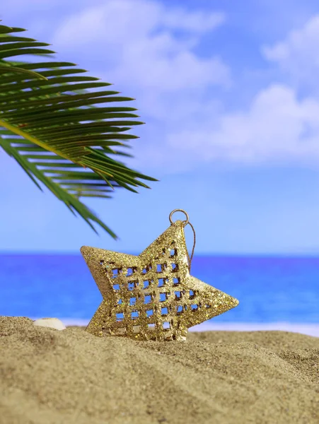 Xmas summer holidays concept. Christmas ornament on a sandy beach with palm tree, blue sky and sea background. Vertical photo