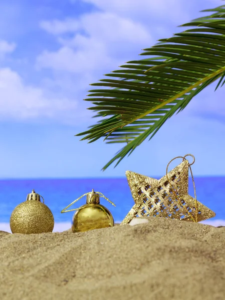 Xmas summer holidays concept. Christmas ornaments on a sandy beach with palm tree, blue sky and sea background. Vertical photo