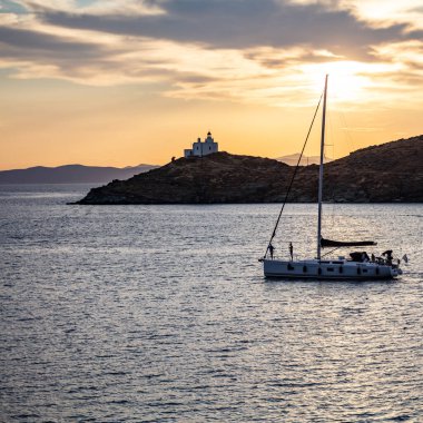 Sakin deniz ve turuncu gökyüzü arka planındaki yelkenli, Yunanistan 'daki Ege Akdeniz' de gün batımında denize açılacak. Kea adasındaki deniz feneri.