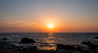 Kayalık bir sahil üzerinde gün batımı turuncu rengi. Ege Denizi Yunanistan 'da dramatik büyülü gündoğumu deniz manzarası. Ada silueti ve dalgalı suda altın yansımalar