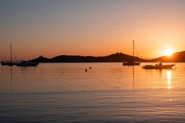 Kea Adası, Yunanistan. Deniz suyunda gün batımı turuncu rengi. Vourkari limanında demirli tekneler, dalgalı sularda altın yansımalar, Ege Akdeniz.
