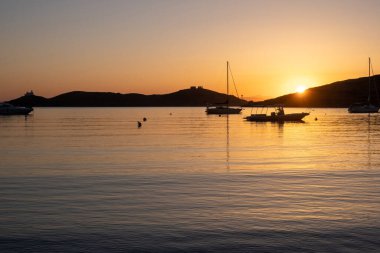 Kea Adası, Yunanistan. Deniz suyunda gün batımı turuncu rengi. Vourkari limanında demirli tekneler, dalgalı sularda altın yansımalar, Ege Akdeniz.