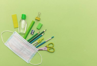 Okula, koronavirüs günlerine. Tıbbi koruyucu maske, el dezenfektanı jeli ve okul malzemeleri pastel yeşil arka plan, üst görünüm, şablon, kopya spac
