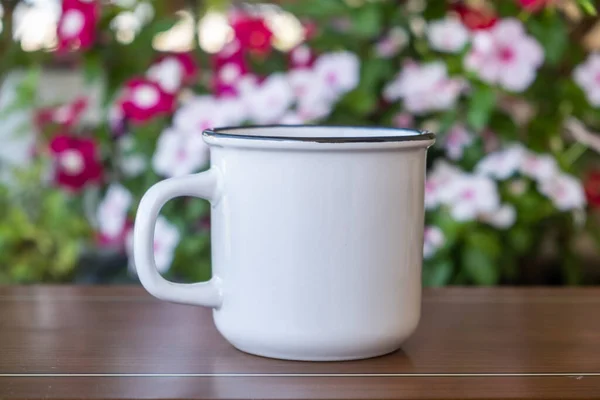 コーヒーカップは庭のテーブルの上でモックアップ 熱い飲み物のためのハンドルを持つ白い色の空白のマグカップは 背景の花を咲かせます ブランディングテンプレート — ストック写真