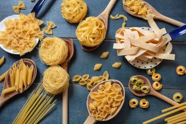 Makarna pişirme konsepti. Çiğ makarna çeşitli şekiller ve kaşıklar düz uzandı mavi ahşap masa arkaplanı, üst görünüm