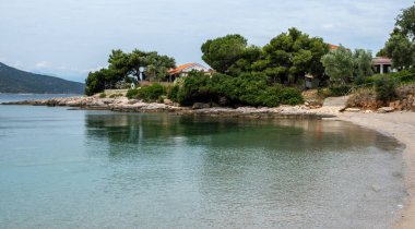 Yunanistan 'ın Batı Attica kentindeki huzurlu Porto Germeno plajı, sakin turkuaz sular, çam kaplı uçurumlar ve uzak dağlar, parçalı bulutlu yaz gökyüzü