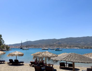 Yunanistan 'ın Poros Adası' ndaki Mikro Neorio Körfezi 'nde yaz sahnesi. Saman şemsiyeler, şezlonglar, turkuaz deniz, demirli yelkenliler ve uzak Poros şehir manzarası.