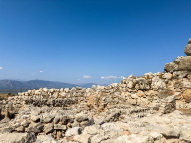 Yunanistan 'daki Mycenae arkeolojik sahasında, açık mavi yaz gökyüzünün altında, arka plandaki uzak dağlar ve parlak güneş ışığının altında antik taş duvar kalıntıları bulunuyor.