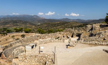 Yunanistan 'daki Miken arkeoloji sahasındaki antik taş kalıntıları, güçlendirilmiş duvarlar, dairesel yapılar ve açık mavi gökyüzü altındaki dağ manzarası
