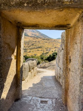 Yunanistan 'daki Miken arkeoloji sahasındaki antik bir taş geçitten bakıldığında, engebeli tepeler ve güneş ışığı altındaki altın yaz manzaraları görülüyor.