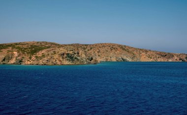 Yunan kıyı şeridi boyunca uzanan sakin derin mavi denizin üzerinde engebeli bir kayalık yükseliyor, berrak gün ışığında sakin bir ufuk ve yumuşak doğal tonla yakalanıyor..