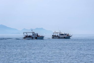 Korfu, Yunanistan, 5 Haziran 2025. Turistlerle dolu iki tur teknesi yumuşak ışık altında sisli dağ siluetleriyle durgun mavi denizi aşıyor..