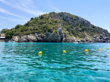 Korfu, Yunanistan 'da turkuaz deniz suyuyla kayalık kıyı şeridi. Güneşli bir yaz günü, berrak gökyüzü, yemyeşil ve sudaki sakin yansımalar