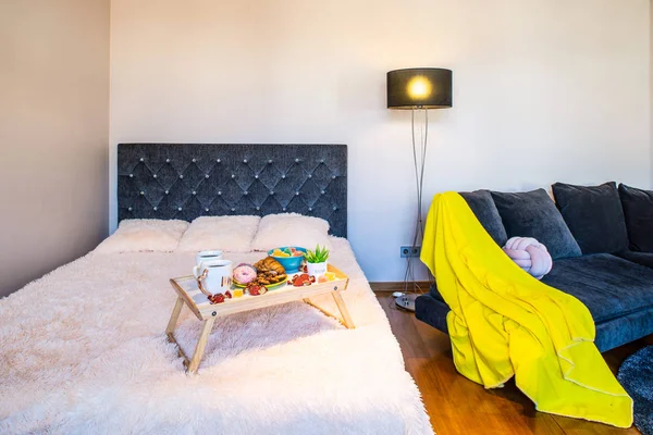 Covered bed and sofa in light studio apartment. Food on the tray.