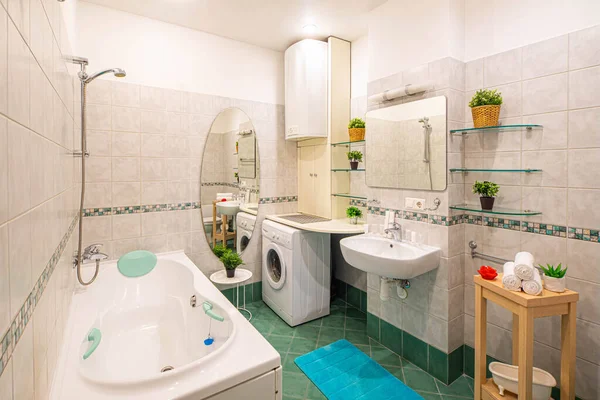 Light interior of modern spacious bathroom. White sink and mirror. Flowers on shelves. Large bath. Washing machine.