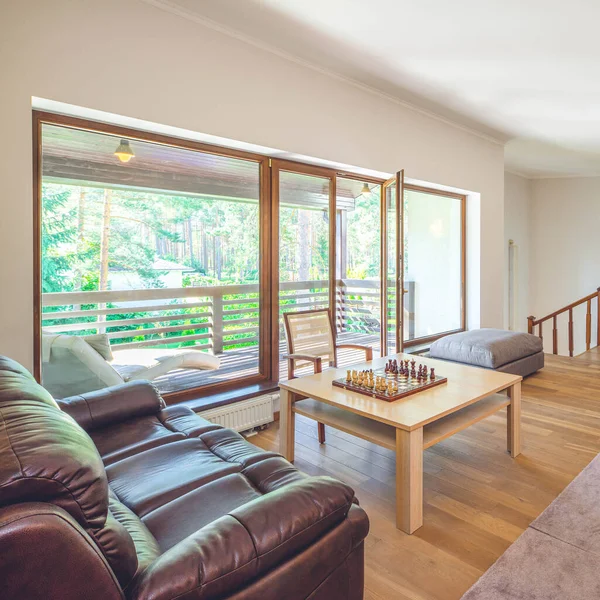 Modern interior of hall in luxury private house. Chess on table. Wooden chair. Leather couch. Glass door to terrace.