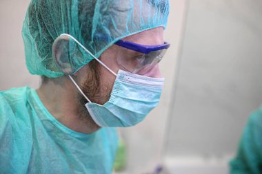 Gözlüklü ve koruyucu maskeli bir cerrah ameliyathanede çalışıyor.
