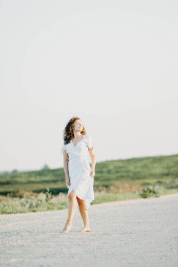 Kıvırcık genç kadın, gün batımında kumlu bir yolda mavi bir elbiseyle romantik bir şekilde dans ediyor..