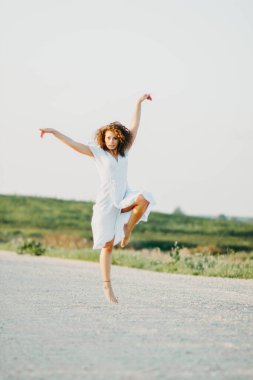 Kıvırcık genç kadın, gün batımında kumlu bir yolda mavi bir elbiseyle romantik bir şekilde dans ediyor..