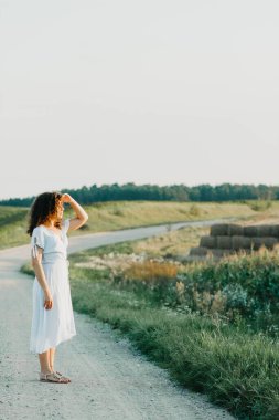 Kıvırcık genç kadın, gün batımında kumlu bir yolda mavi bir elbiseyle romantik bir şekilde dans ediyor..