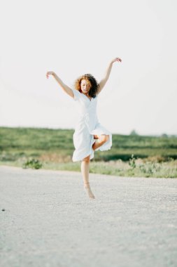 Kıvırcık genç kadın, gün batımında kumlu bir yolda mavi bir elbiseyle romantik bir şekilde dans ediyor..