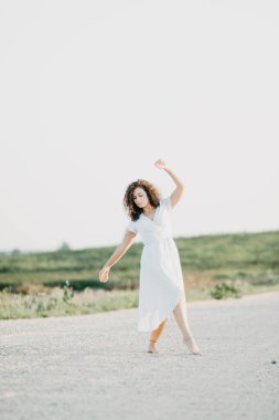 Kıvırcık genç kadın, gün batımında kumlu bir yolda mavi bir elbiseyle romantik bir şekilde dans ediyor..