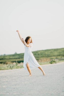 Kıvırcık genç kadın, gün batımında kumlu bir yolda mavi bir elbiseyle romantik bir şekilde dans ediyor..