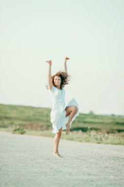 Kıvırcık genç kadın, gün batımında kumlu bir yolda mavi bir elbiseyle romantik bir şekilde dans ediyor..