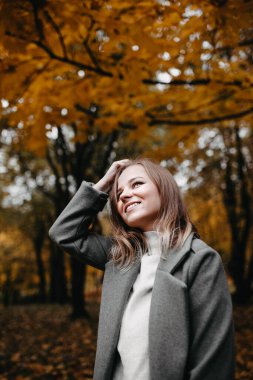 Gri montlu genç bir kadın sonbahar ormanında yürüyor. 