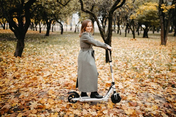 Woman in autumn coat on an electric scooter in an autumn park. Riding on electric vehicle in cold weather