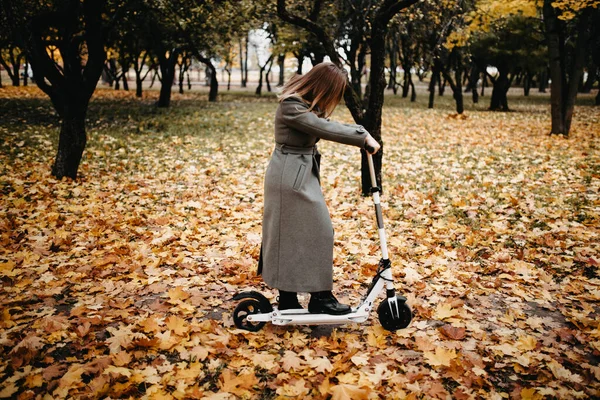 在秋天的公园里 穿着秋天外套的女人坐在电动车上 寒冷天气下骑电动车 — 图库照片