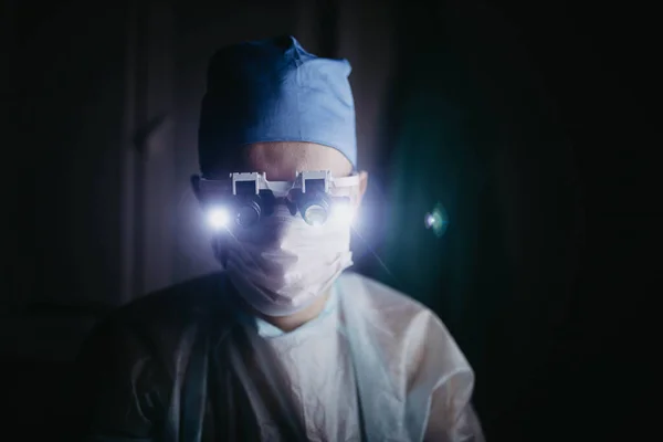 両眼拡大鏡を身に着けている外科医は暗い手術室で患者に動作します — ストック写真