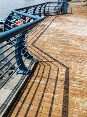 Bu şehir manzarası, korkulukların ışığı ve gölgesi tarafından oluşturulan ahşap iskelenin geometrik desenini yansıtıyor. Bu kompozisyon mimari tasarım ve doğa arasındaki etkileşimi vurgular.