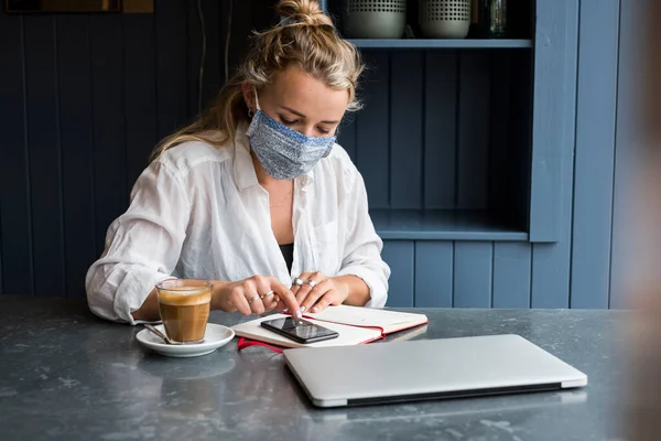 Donna che indossa maschera viso da sola a un tavolo da caffè con un computer portatile e notebook che lavora da remoto. — Foto stock