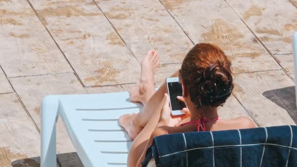 女人在游泳池旁的椅子上享受日光浴和使用手机 — 图库视频影像