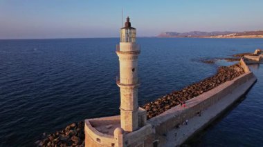 Kayalık sahilde yükselen bir deniz feneri günbatımında gemilere rehberlik ediyor. Dalgalar kıyıya vururken gökyüzü akşam renklerine bürünüyor..