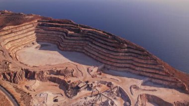 Açık çukur madeninde deniz kenarında farklı katmanlı kaya oluşumları bulunur. Günbatımının altın renklerini yansıtır. Doğanın ve madenciliğin bir karışımını vurgular..