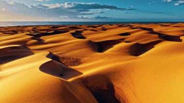 Altın kum tepeleri, batan güneşin sıcak renkleriyle aydınlanan Gran Canaria manzarası boyunca uzanır ve deniz kenarında nefes kesen doğal bir tuval oluşturur..