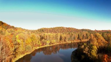 Berrak bir mavi gökyüzü altında parlak turuncu, sarı ve yeşil yapraklar sergilenen sakin bir nehir, sonbahar boyunca canlı bir ormanın içinden akar..