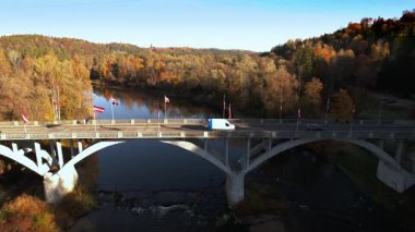 Bir kamyon köprüden geçiyor, sakin bir nehre bakıyor, canlı sonbahar ağaçları kıyıları kaplıyor, güzel bir sonbahar manzarası yaratıyor..