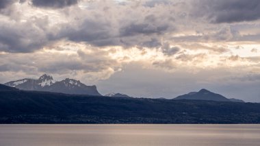 Geniş panoramik açıdan Geneva Gölü 'nün bulutlu bir gün batımının altında gökyüzünde parlayan ışık ve uzaktaki Alpler.